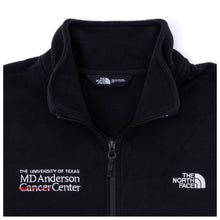 Closeup of a black North Face fleece jacket featuring the white MD Anderson logo on the right side of the chest and the white North Face logo on the left side of the chest, featuring a full zipper and front zippered-pockets.