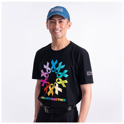 MD Anderson employee wearing a black t-shirt adorned with colorful cancer ribbons forming a circle and the phrase 'Stronger Together' in various colors, with the white MD Anderson logo featured on the sleeve, and also wearing the denim blue cap featuring the white MD Anderson logo. 
